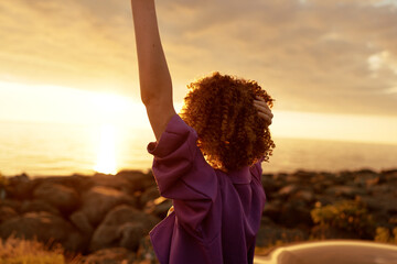 Obraz premium Rear view of a woman with curly hair raising her arm at sunset by the sea, wearing a purple top, capturing a carefree lifestyle moment and warm golden light.