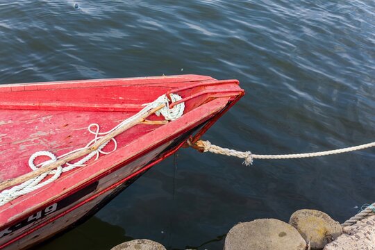 A top-down view of a weathered red wooden boat bow, tied with thick white ropes to a rustic pier made of wood posts and stone, floating on calm blue water