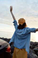 Woman stands by the coast with arms raised, wearing a blue shirt and hat, enjoying sunset, exuding outdoor lifestyle, freedom and joy by the sea © SHOTPRIME STUDIO