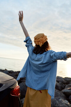 Woman stands by the coast with arms raised, wearing a blue shirt and hat, enjoying sunset, exuding outdoor lifestyle, freedom and joy by the sea