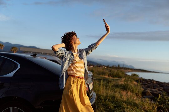 Cheerful woman outdoors, sunlit moment beside a car, coastal background, relaxed posture, striped top, yellow skirt, gentle breeze, natural colors, vibrant mood and joyful energy tonight