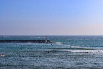 Obraz premium Lighthouse at the End of a Coastal Pier with Surfers Waiting for Waves