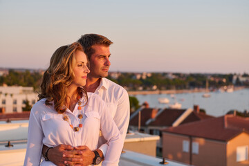 Obraz premium Romantic Couple Embracing on Rooftop Balcony Overlooking Seaside Town at Sunset