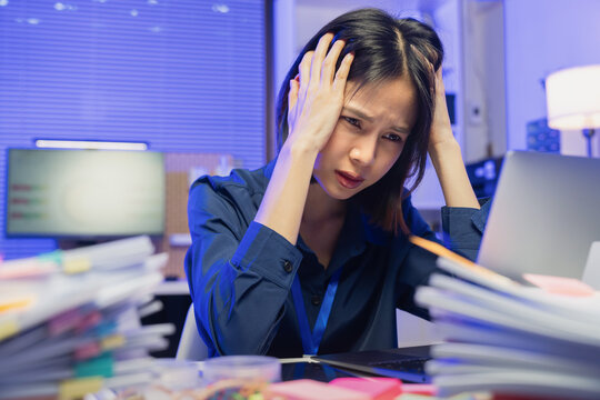 Stressed young Asian businesswoman holding head while working on laptop in office, burnout and work pressure concept.