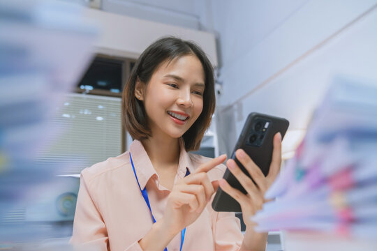 Smiling young Asian businesswoman using smartphone while working in office, mobile communication and business technology concept.