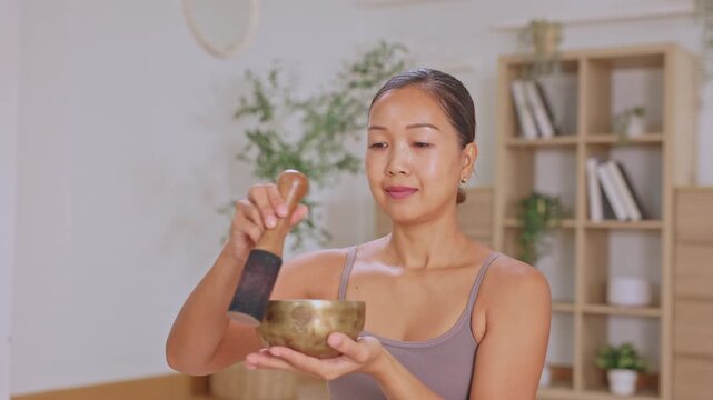 Asian woman singing bowl meditation wellness calm indoor relaxation sound therapy mindful home practice with metal bowl wooden mallet peaceful self care cozy room plant shelf