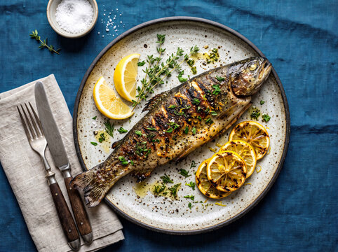 Whole grilled trout with lemon slices and fresh thyme served on plate with blue linen cloth.