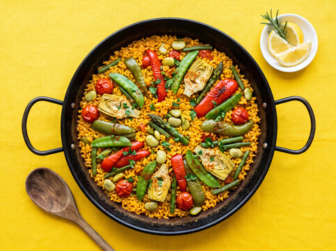 Vegetable paella in pan with artichokes, peppers, peas, asparagus and lemon slices.