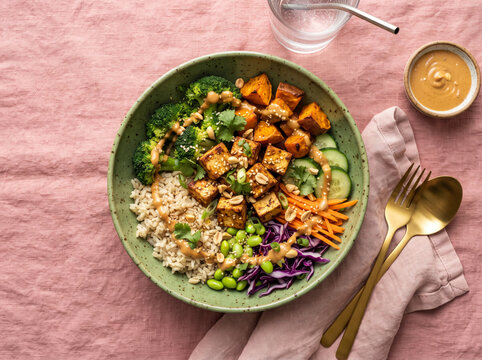Healthy vegan bowl with crispy tofu, roasted sweet potatoes, and peanut dressing on pink background.