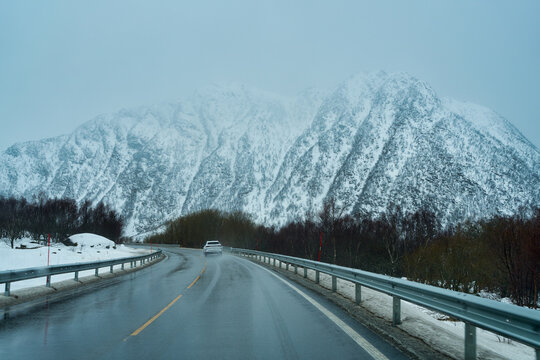 POV driving a car on a wet winding highway following a white vehicle toward massive snow-covered mountains shrouded in thick mist and fog. Scenic winter road trip adventure in Lofoten, Norway, Scandin