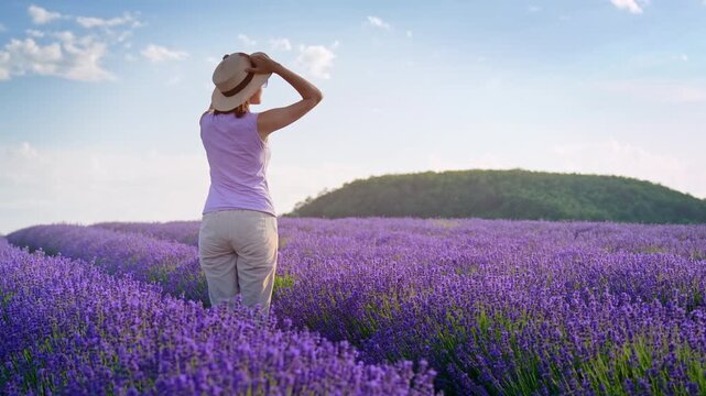 Young woman in a straw hat raising her arms and enjoying freedom in a blooming lavender field at sunset. Handheld motion footage for summer travel and wellness video concept.