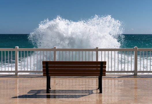 Empty bench faces ocean where powerful wave erupts behind railing
