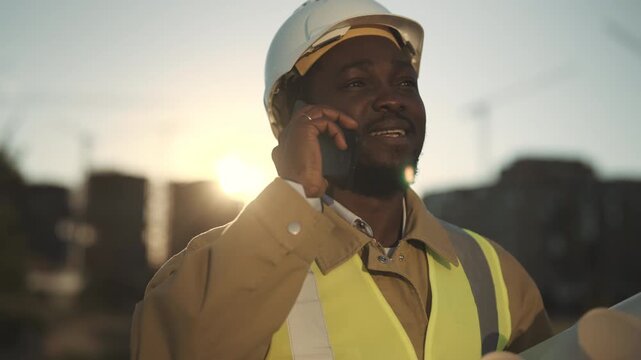 Professional foreman talking by phone in construction site, portrait in sunset. Engineer or architect calling to investors and reporting about works progress, african builder with white hard hat