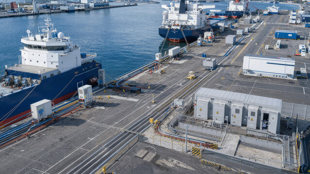 Ships dock at a busy port with containers and buildings near the sea during a sunny day with clear skies