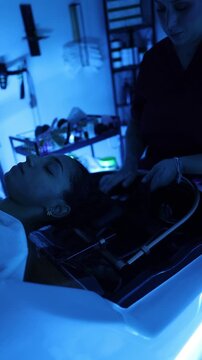Young woman relaxing while a professional hairdresser washes her long dark hair in a modern salon sink, preparing her for a rejuvenating scalp and hair care treatment under soothing blue light