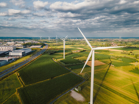 Aerial drone view of wind turbines standing among green patchwork farm fields beside a motorway under a cloudy summer sky, a scene of renewable wind energy and sustainability.