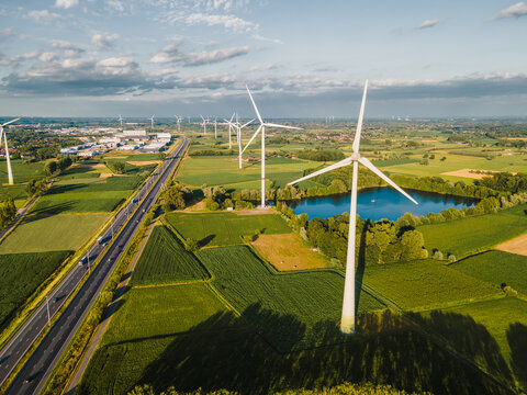 Aerial view of wind turbines among lush green farm fields with a blue pond and a highway in warm evening light, a landscape of renewable green energy.