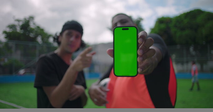 Two Hispanic men on soccer field smiling as one holds smartphone with green screen toward camera while friend points excitedly, mockup concept for mobile app, social media, outdoor technology