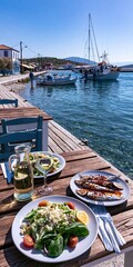 Fototapeta premium Enjoying fresh seafood and greek salad on a wooden pier overlooking a harbor with fishing boats and a sailboat