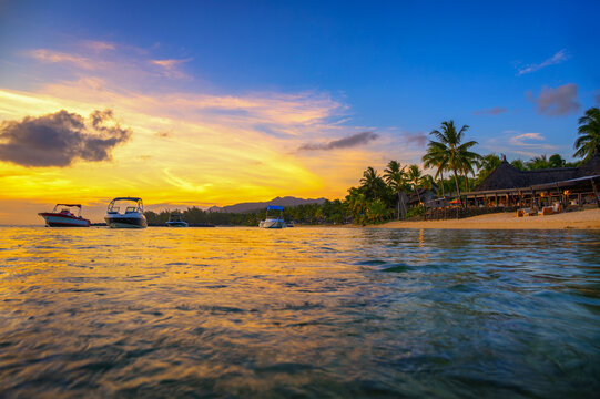 Colorful sunset over Bel Ombre beach in Mauritius with calm water, anchored boats, palm lined resorts, and distant mountains beneath a glowing tropical sky.