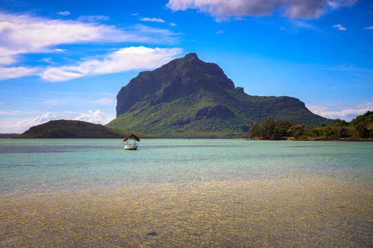 Small boat floats in a turquoise lagoon below Le Morne Brabant, a UNESCO World Heritage mountain on Mauritius, with tropical shoreline and bright clouds.