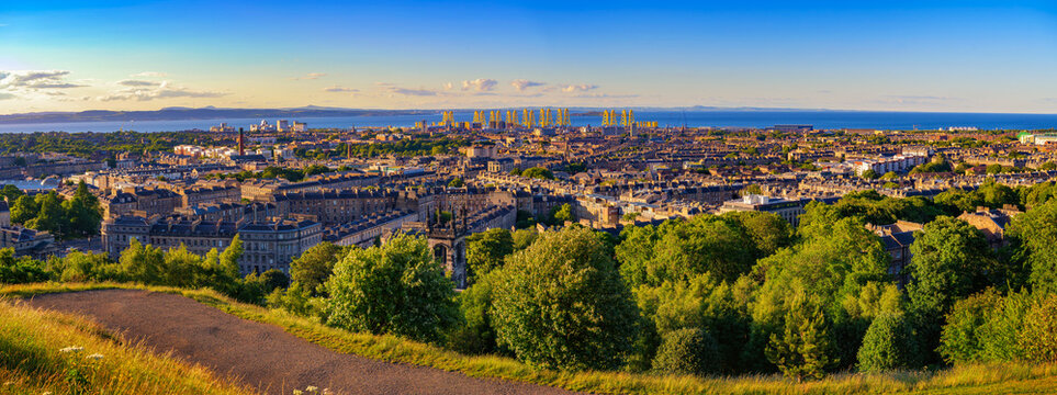 Panoramic sunset view from Calton Hill over Edinburgh rooftops, Leith Docks, and the Firth of Forth, with green hillside foreground and warm evening light in Scotland, United Kingdom.