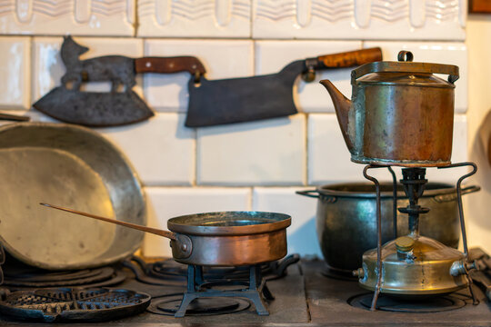 Kitchen still life featuring an antique copper kettle on an old spirit stove, a small copper saucepan on a cast iron trivet, and vintage butcher knives and metal cookware hanging against a white tiled
