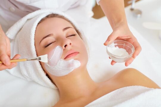 Young woman receiving a professional facial mask treatment at a beauty spa, relaxing with closed eyes while a cosmetologist applies moisturizing skincare cream.