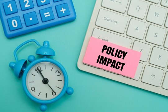 Pink sticky note with "POLICY IMPACT" text on a keyboard, next to a blue alarm clock and calculator against coloured background. Concept of business regulation change and economic time management