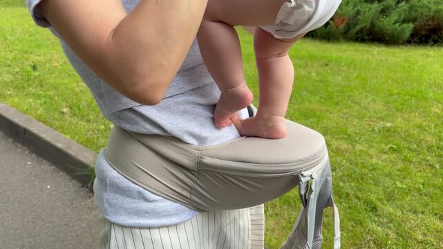 Close-up of a baby standing and bouncing on a hip seat carrier held by a mother outdoors. Active parenting, early childhood development, and modern baby gear.