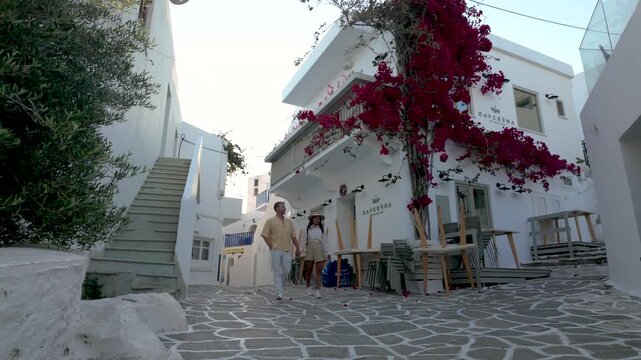 A couple wanders through the captivating streets of Naousa, Paros, Greece, where quaint buildings and vibrant bougainvillea create a picturesque holiday experience. Enjoy the Cycladic beauty