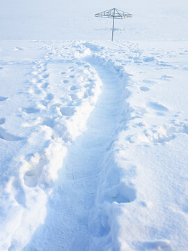 Snow path winds across deep drifts toward empty umbrella frame. Winter landscape stretches under pale light, while snow covers quiet ground with crisp footprints and calm atmosphere