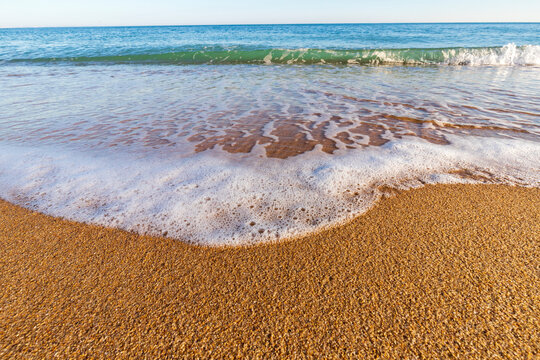Close-up of soft sea wave foam washing over a golden sandy beach on a sunny dayClose-up of soft sea wave foam washing over a golden sandy beach on a sunny day