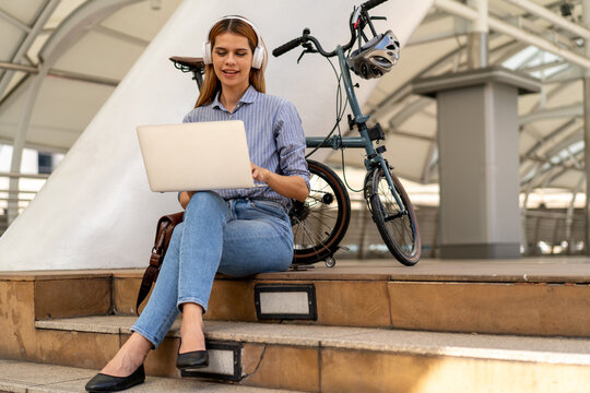 Urban Commute: A young woman wearing headphones works on her laptop while sitting on outdoor steps next to her foldable bicycle, blending technology and active urban lifestyle.