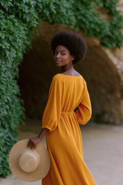 Graceful woman in mustard dress holding a straw hat beside an ivy covered stone archway for fashion portrait and vertical background design
