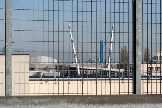 Recinzione metallica con motivo a griglia in primo piano, che offre la vista di una struttura al di l&agrave;. La struttura ricorda un ponte sospeso o un elemento architettonico moderno.