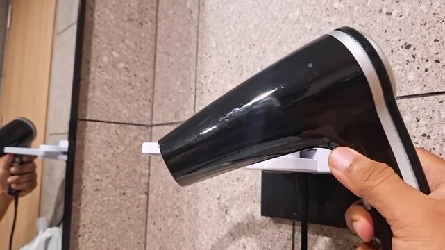 close-up shot showing a hand picking up a black handheld hair dryer from a wall mount next to a mirror, turning it on briefly to demonstrate the airflow, and then placing it back.