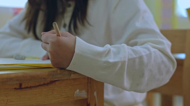 Student hand holding pencil over paper at desk in classroom, writing study education scene with white sweater sleeve, close view of young learner in school setting