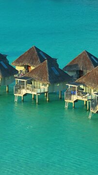 Vertical Screen: Bora Bora overwater bungalows with traditional thatched roofs stand on stilts above crystal clear turquoise lagoon waters with private decks and ladders for ocean access.