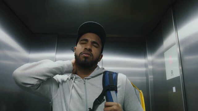 Tired Delivery Worker Wearing Cap And Backpack Adjusting Hoodie Inside Elevator, Sweating After Work