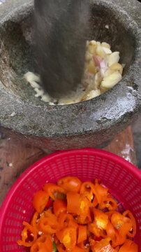 Crushing garlic in a traditional stone mortar and pestle with sliced red chili peppers in a basket foreground.