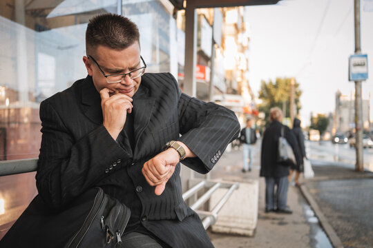 Businessman sitting at a bus stop, looking anxiously at his watch while waiting for transport. Running late for work, time pressure, and urban morning stress.