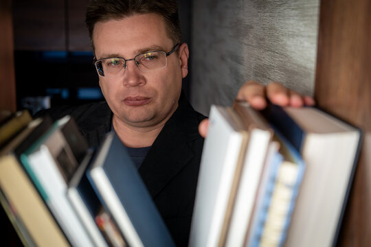 Serious man with glasses selecting a book from a wooden bookshelf, looking focused in a library or home study setting.