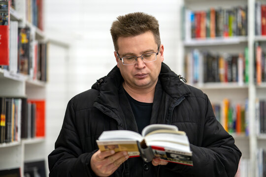 Man with glasses reading an open book while standing in the aisle of a bright bookstore with white shelves.