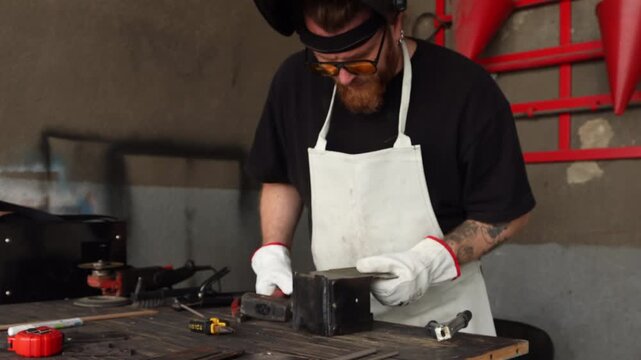 Bearded craftsman in protective apron and safety glasses shaping metal with hammer