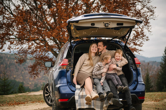 A joyful family, including a father, mother, son, and daughter, sharing a candid, happy moment while sitting in the open trunk of a blue SUV during an autumn mountain trip.