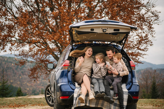 A candid family portrait of a mother, father, son, and daughter laughing and interacting while sitting in the open trunk of a blue SUV during an autumn picnic in the mountains.