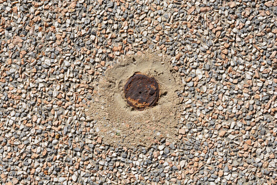 Full frame view of an old rusty metal pipe cap embedded in a smooth concrete circle surrounded by exposed aggregate terrazzo wall texture

