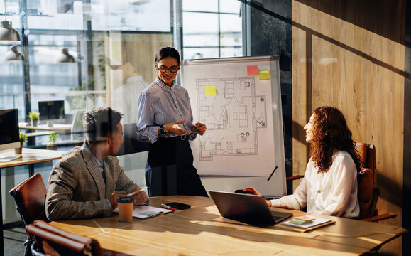 A smiling woman, wearing glasses and a striped shirt, presents a hand drawn floor plan on a flipchart easel, gesturing toward the diagram while two seated colleagues, a man in a blazer and a curly