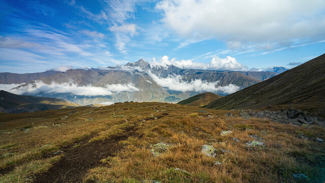 golden grass ridge overlooks a cloud-filled valley beneath snow-capped Mount Kazbek under a blue sky, Georgia.